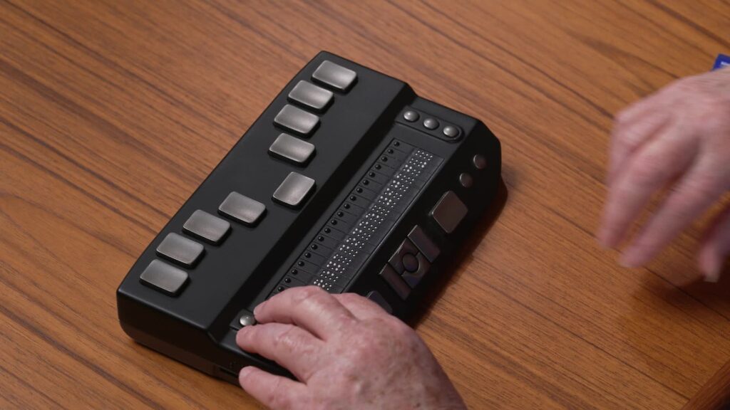 Picture of the Zoomax e-reader, showing the top of the reader with a typical Perkins keyboard layout.  A hand is shown on the 20-cell braille display. The device is 8 inches wide, 5 inches deep, and 1.5 inches tall.